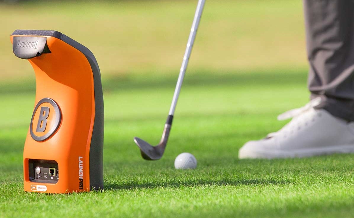 Golfer using launch monitor on green grass.