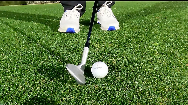 Golf club poised above white golf ball on grass.