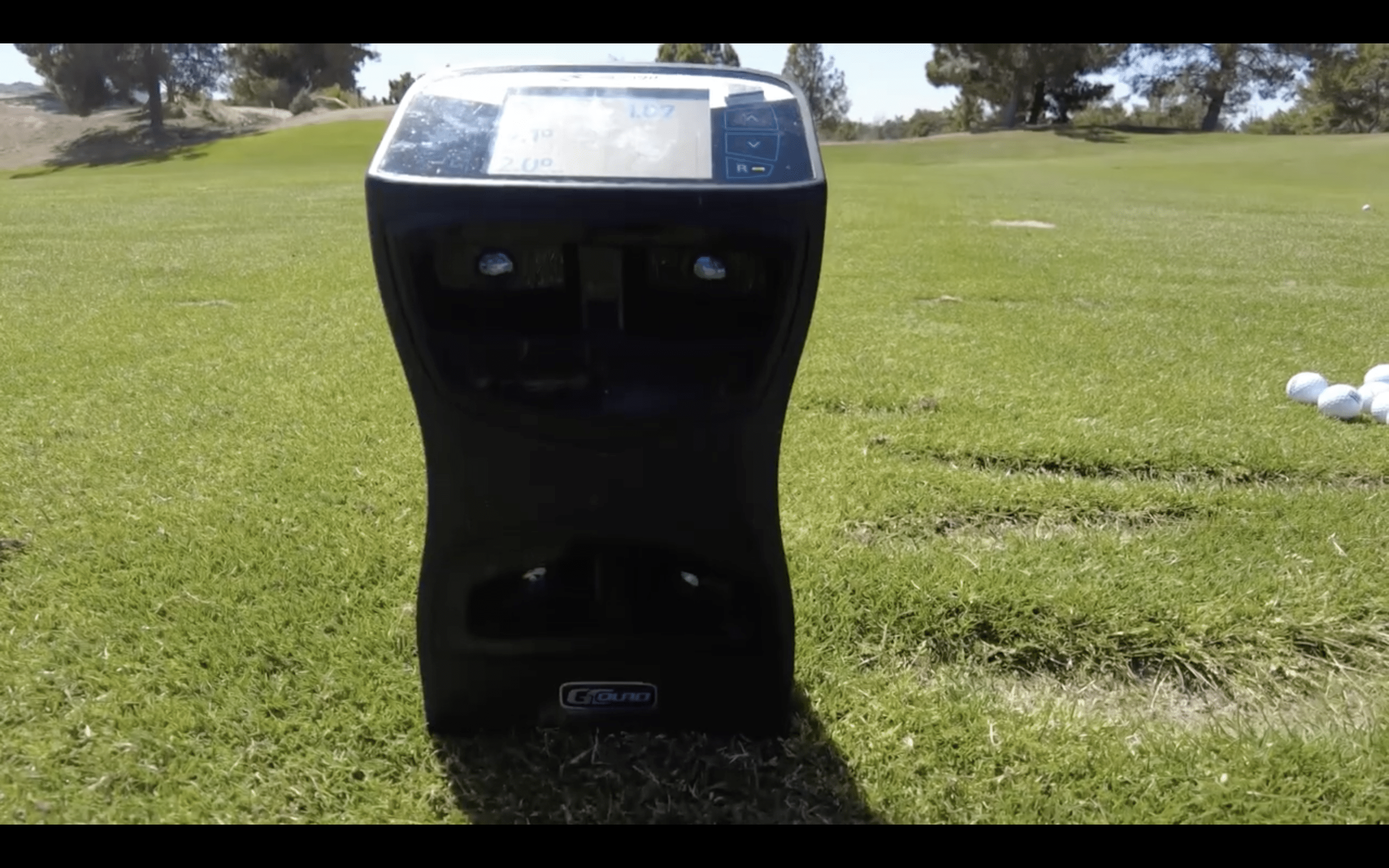 Golf launch monitor on grass with golf balls nearby.