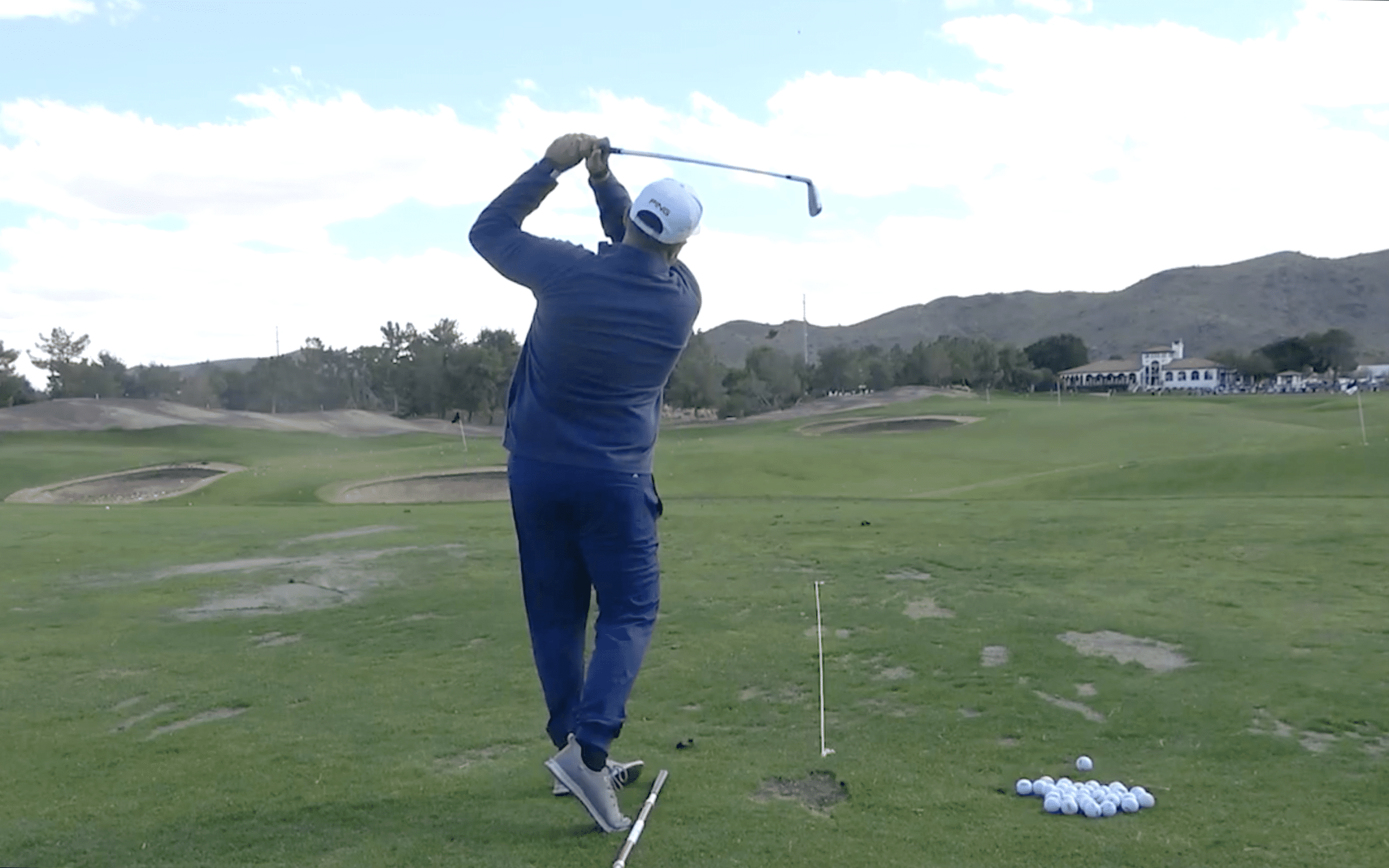 Golfer swings on practice range with mountains behind.
